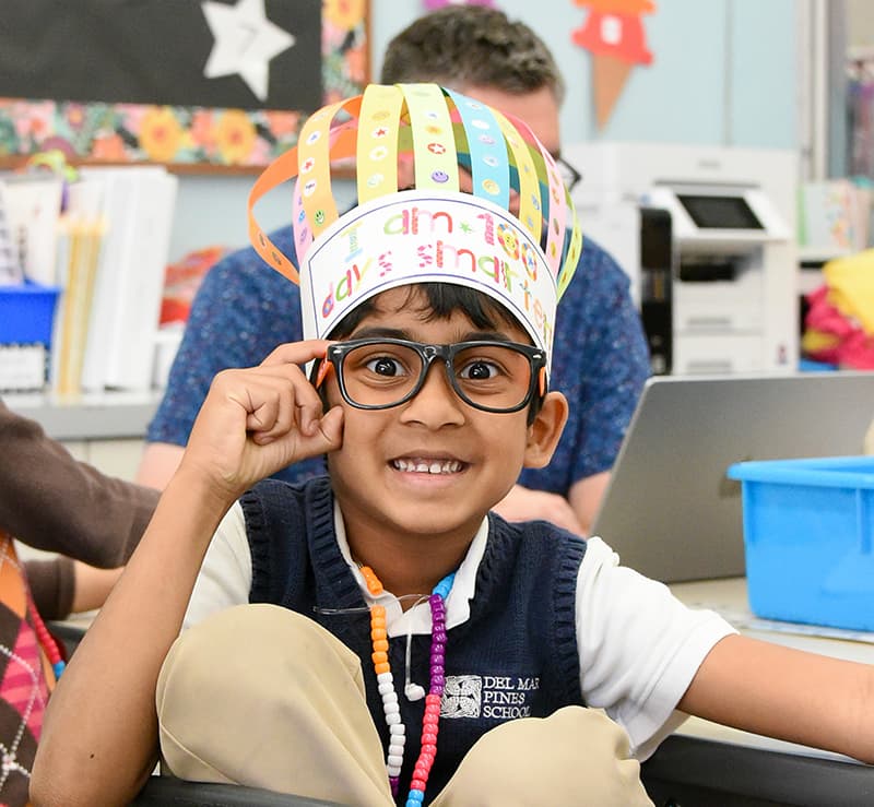 Student wearing a paper crown and glasses, smiling in class