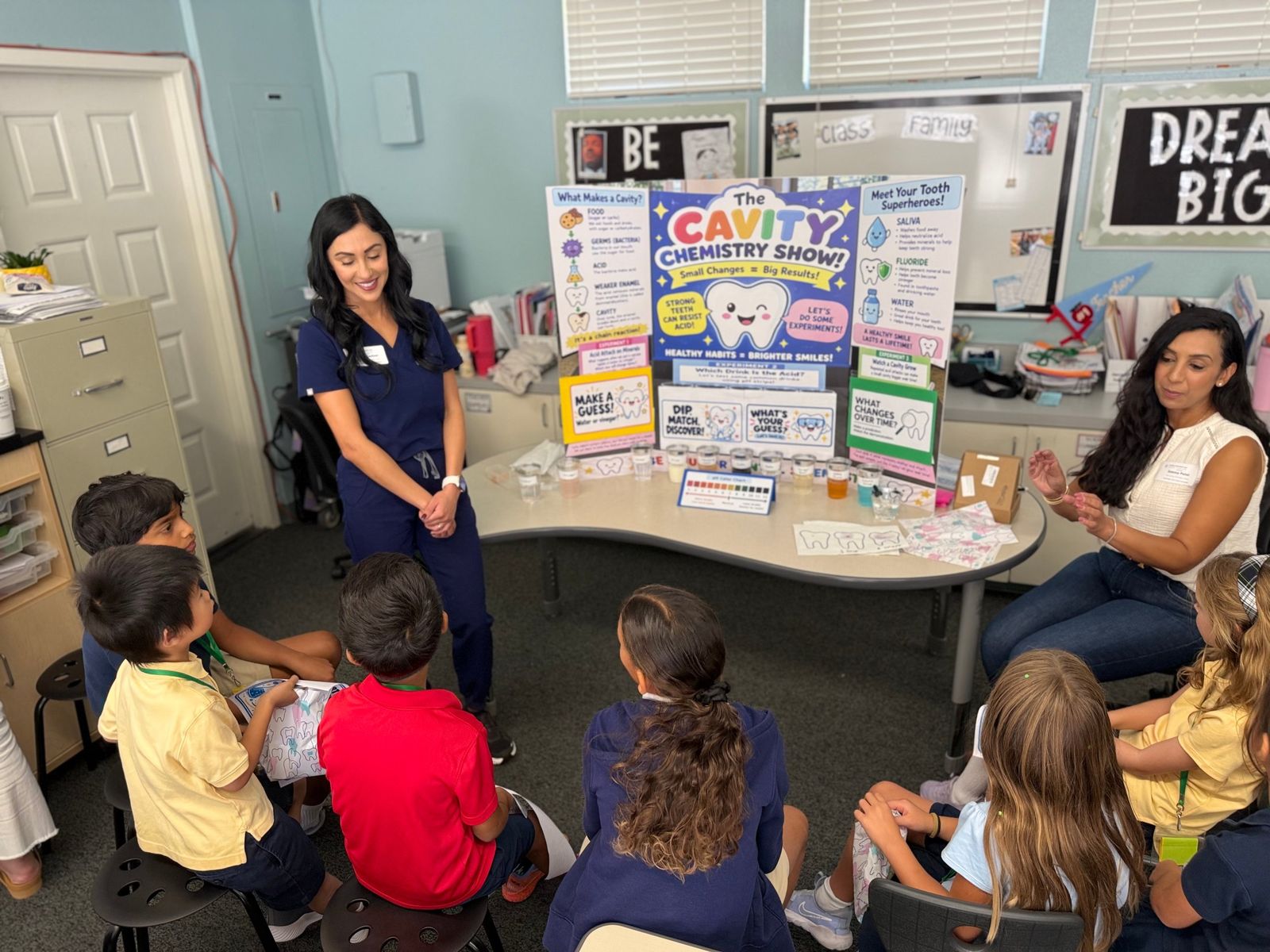 A parent presenter in scrubs leads students through The Cavity Chemistry Show, with a display board and pH test cups on the table.